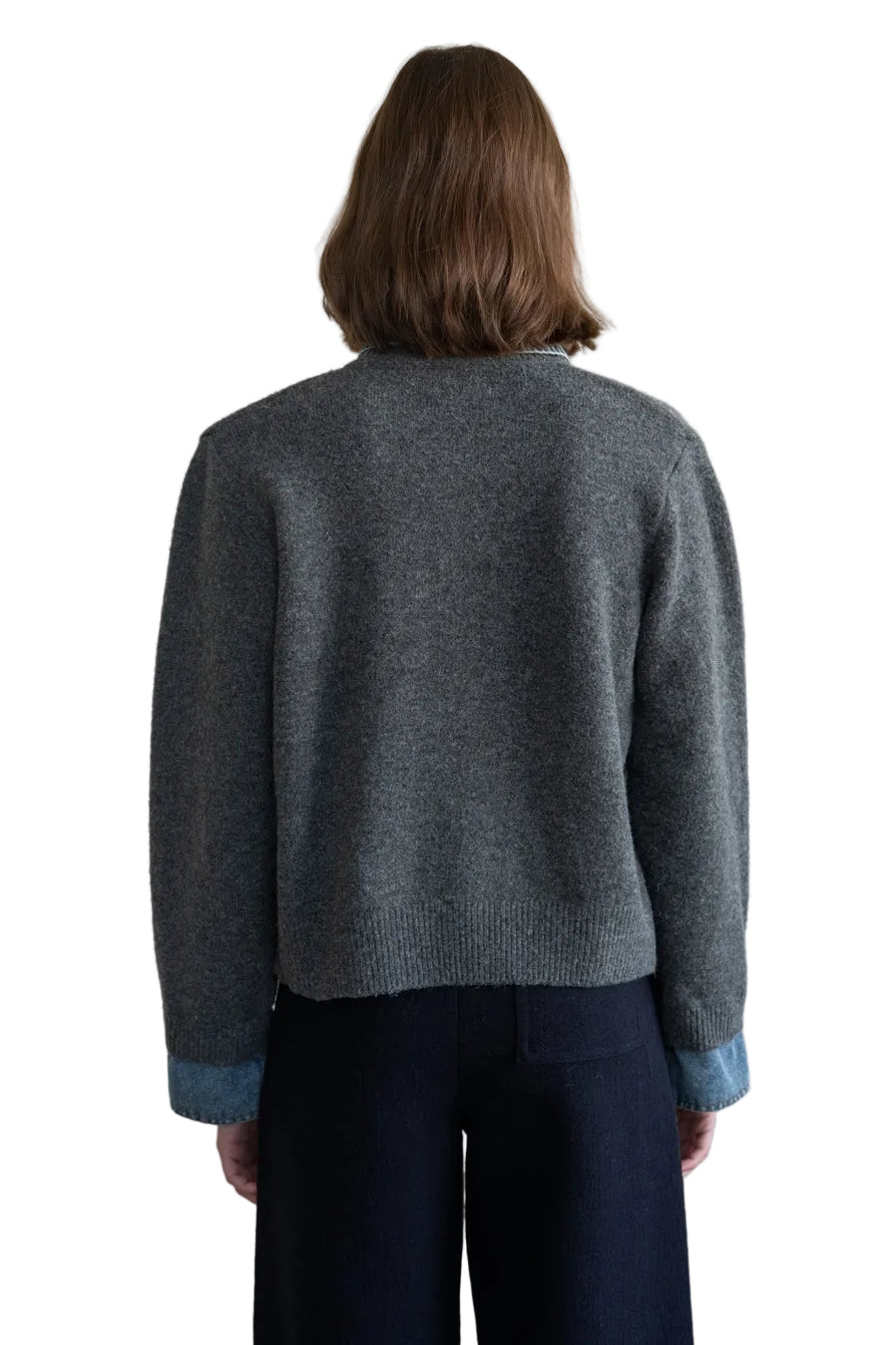 A person shown from the back wearing a dark gray knit sweater with light blue ribbed cuffs and dark navy pants, standing against a plain light beige wall.